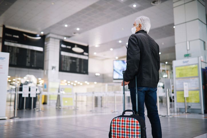 Senior man traveling in the times of the coronavirus pandemic. He is carrying a roller bag on the empty airport.