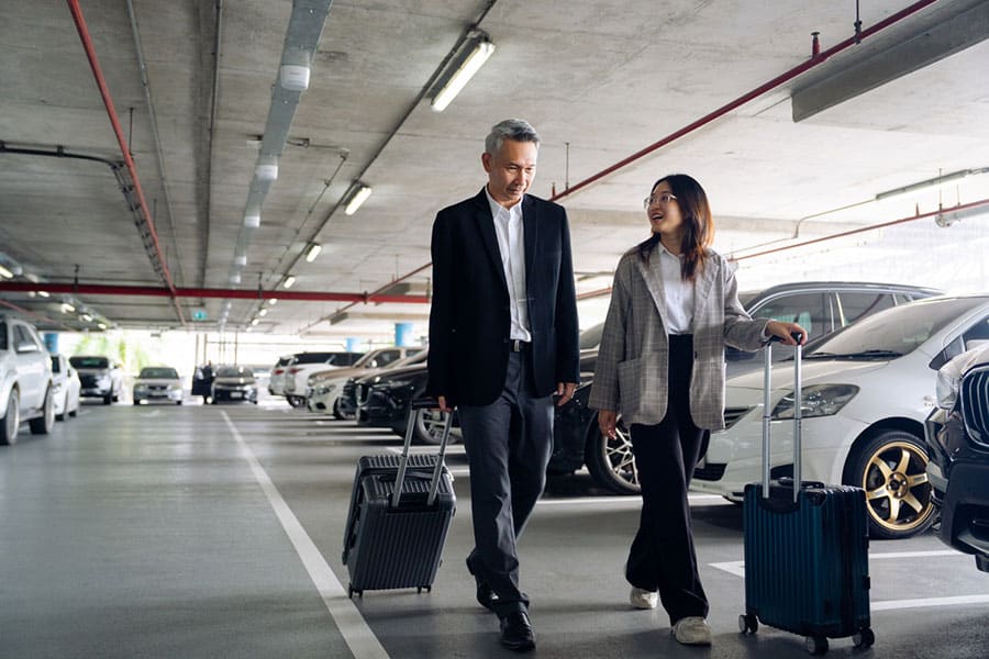 Two travelers walk through an airport parking garage, each pulling a suitcase. Dressed in business attire, they appear to be smiling and talking to each other as they navigate their airport parking options, surrounded by parked cars under a concrete ceiling.