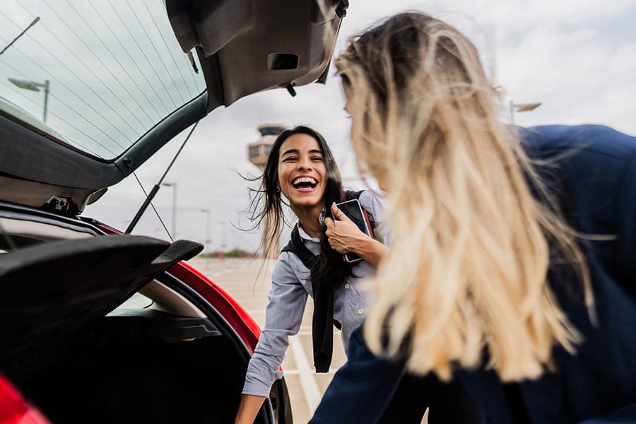 Two women smiling and unloading items from the trunk of a red car in an airport parking lot on a cloudy day, capturing the convenience and joy that many travelers experience when choosing flexible airport parking options.