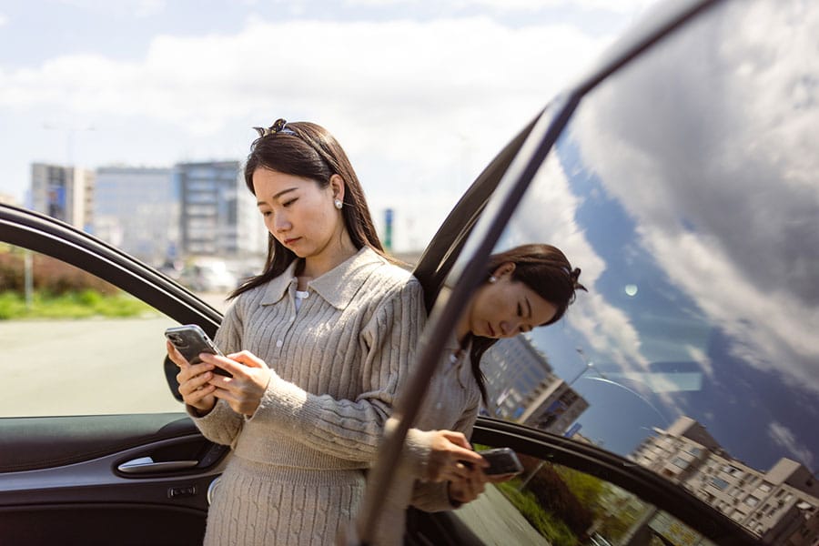A woman stands outside a car with the door open, looking at her phone—perhaps checking DTW Parking options. Her reflection appears in the window, with buildings and a blue sky with clouds in the background.