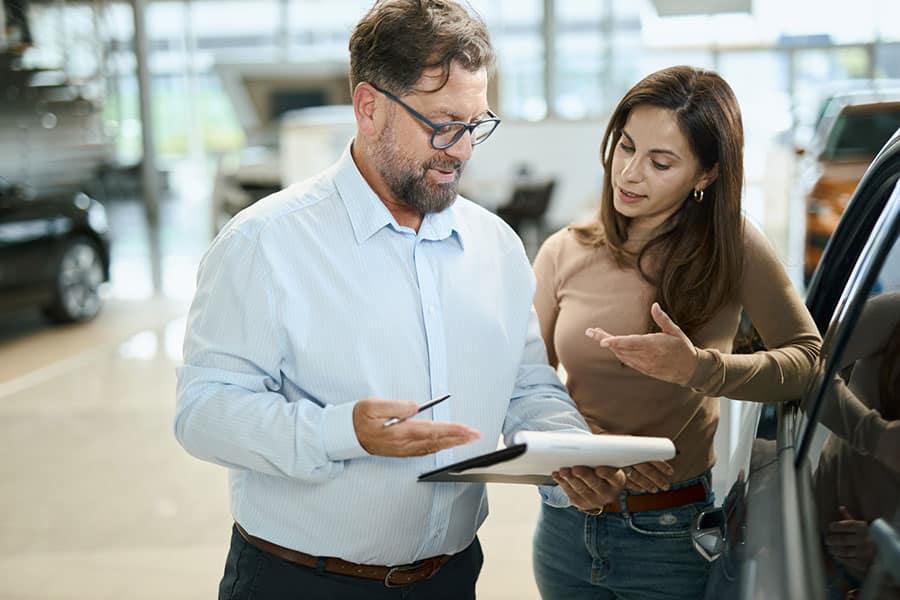 A man holding a clipboard discusses car inspection paperwork with a woman next to a car in a dealership showroom. Both appear focused and engaged in conversation.