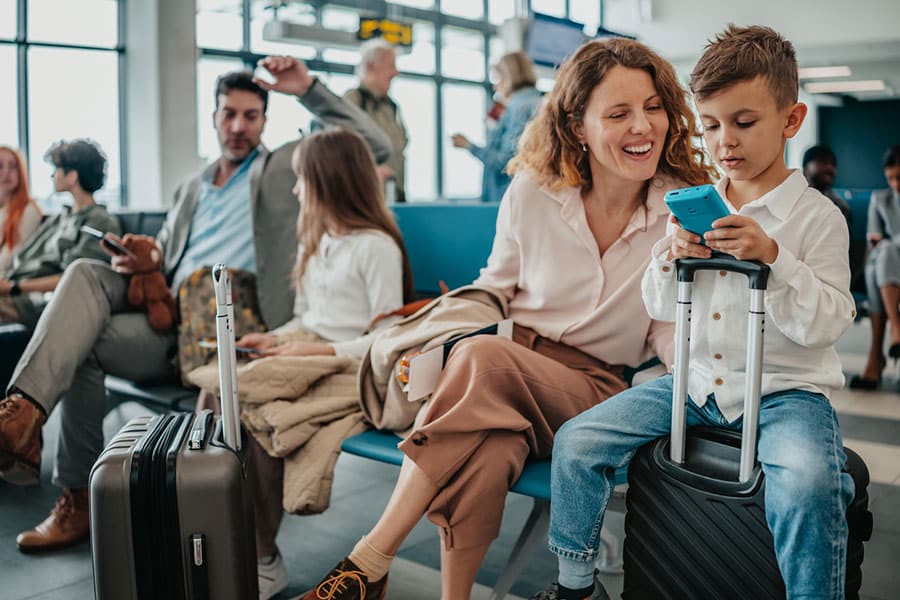 A woman and boy sit in a Detroit airport waiting area. The boy uses a smartphone while sitting on a suitcase, surrounded by fellow travelers. Other passengers, including a man and girl, are visible in the background, possibly awaiting DTW Valet Parking services.