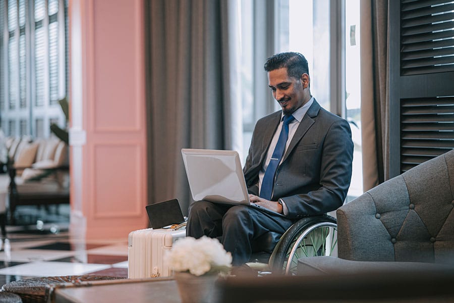 A smiling man in a business suit works on a laptop while seated in his wheelchair in a hotel lobby, as if preparing for a traveler personality test.