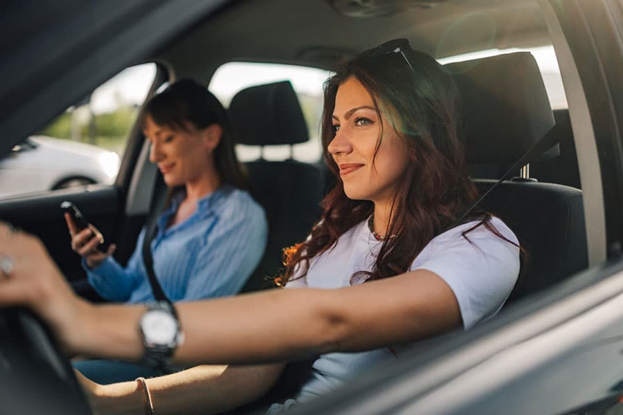 A woman driving a car for airport transportation, with a passenger looking at a phone in the seat behind her.